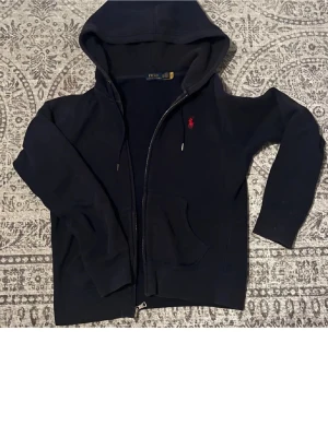 Svart hoodie från Polo Ralph Lauren - Svart hoodie med dragkedja från Polo Ralph Lauren. Klassisk design med huva och fickor framtill. Ikoniska röda Polo-loggan broderad på bröstet. Tillverkad i mjukt bomullsmaterial som är skönt att bära. Perfekt för en avslappnad och stilren look.