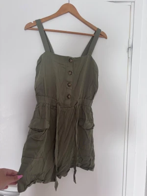 Olivgrön kort jumpsuit med knappar - Snygg olivgrön jumpsuit med breda axelband, knappar framtill och resår i midjan. Modellen har två stora fickor på sidorna och korta ben. Tillverkad i ett lätt, mjukt tyg som ger en avslappnad look. Perfekt för varma dagar och enkel att styla.