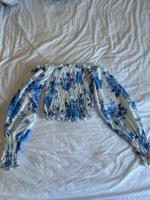 Blåvit offshoulder blus med blommor - Säljer en snygg offshoulder blus med blåa blommor på vit bakgrund. Blusen har smockad kropp och långa puffiga ärmar med smockade muddar. Materialet är lätt och luftigt, perfekt för sommarens outfits.
