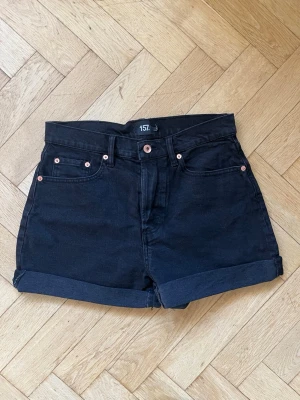 Svarta högmidjade denimshorts  - Säljer ett par svarta högmidjade shorts från 157 med klassisk femficksdesign och uppvikta ben. Shortsens material är denim och de har snygga kopparfärgade nitar och knapp framtill. 