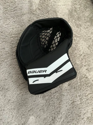 Svart och vit målvaktshandske Bauer - Målvaktshandske från Bauer i svart och vitt med tydlig logotyp. Handsken har ett robust skydd och ett nät för att fånga pucken, perfekt för hockeymålvakter. Materialet är slitstarkt syntet och designen är modern och sportig.