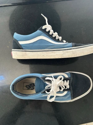 Blå Vans Old Skool sneakers - Säljer ett par klassiska Vans Old Skool sneakers i blått och svart med vit sidostrip och vita snören. Skorna har en låg profil och är tillverkade i canvas och mocka. 