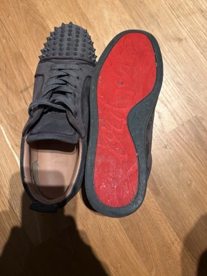 Gråa  spikesneakers från Louboutin - Unika gråa sneakers från Christian Louboutin med mattsvarta nitar på tån och klassisk röd sula. Skorna är låga och har en ovandel i mocka med svarta skosnören. Perfekta för dig som vill sticka ut med en edgy och lyxig stil.