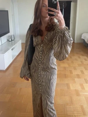 Zebramönstrad långklänning från Adoore - Snygg långklänning från Adoore med zebramönster i beige och grå toner. Klänningen har djup v-ringning, knappar framtill, slits och voluminösa långärmar. Perfekt för dig som vill sticka ut med en trendig look. I storlek xxs