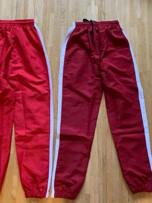 Röda mjukisbyxor med vita ränder - Två par röda mjukisbyxor med vita ränder längs sidorna. Ena paret har snörning i midjan, båda har resår vid benslut och midja. Perfekta för chill eller träning, tillverkade i mjukt material som känns skönt mot huden.