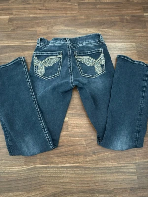 Mörkblå bootcut jeans med brodyr - Snygga mörkblå bootcut jeans med kontrastsömmar och broderade fickor bak i vitt och beige. Jeansen har klassisk femficksmodell och slitningar framtill för en cool look. Perfekta för dig som vill sticka ut lite extra.