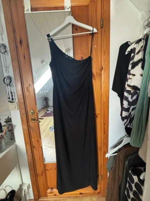 Svart Ralph louren klänning - Säljer en elegant svart långklänning med oneshoulder-design och smal axelband. Klänningen har en stilren och enkel form som ger ett snyggt och modernt intryck. Perfekt för dig som vill sticka ut med en unik siluett. Perfekt för bal!