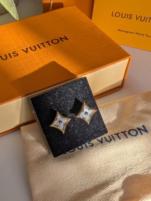 Iouis Vuitton örhänge  - Eleganta stiftörhängen i form av den ikoniska fyrbladiga blomman, presenterade med kompletta tillbehör. Design: Örhängena har en guldfärgad ram med en skimrande vit pärlemorliknande fyllning i mitten. Tillbehör: Levereras med en orange signaturask, en ljus dammpåse i tyg och en orange bärkasse. Detaljer: Inkluderar ett tillhörande infokort märkt "Monogram World Tour". Stil: En sofistikerad och tidlös accessoar som ger en lyxig touch till varje outfit.