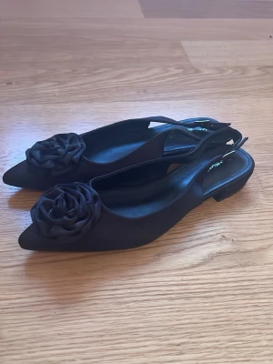 Svarta ballerinaskor med rosdetalj - Stilrena svarta ballerinaskor från Glamorous med spetsig tå och dekorativ ros i tyg framtill. Skorna har öppen häl med justerbar rem och låg klack. Perfekta för dig som vill ha en elegant och trendig look.