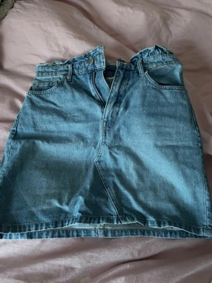 Blå jeanskjol med paperbag-midja - Säljer en blå jeanskjol med paperbag-midja och knäppning fram. Kjolen har fickor både fram och bak samt bälteshällor. Klassisk denimlook som är enkel att styla till olika outfits.