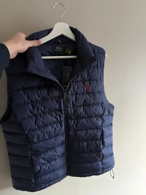 Mörkblå dunväst från Polo Ralph Lauren - Snygg mörkblå dunväst från Polo Ralph Lauren med klassisk quiltad design och röd logga på bröstet. Västen har hög krage, dragkedja framtill och två fickor med dragkedja. Perfekt för lager-på-lager och streetstyle.