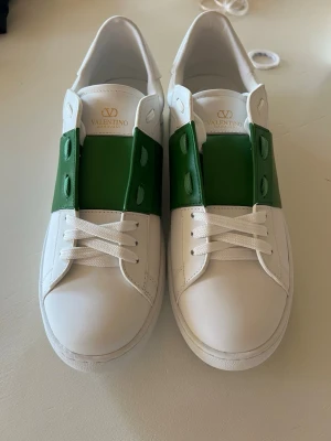 Valentino vita sneakers med grön detalj - Snygga vita sneakers från Valentino med en bred grön läderdetalj över snörningen. Klassisk låg modell med rund tå och vita skosnören. Tillverkade i slätt läder för en clean och exklusiv look. Perfekta för dig som vill sticka ut med stil. Stolek 44 men passar bra för 43 också