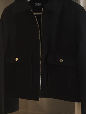 Svart kort jacka från Zara - Svart jacka från Zara med rak och kort passform, dragkedja framtill och två stora fickor med guldfärgade knappar. Jackan har klassisk krage och långa ärmar. Perfekt för att ge en clean och stilren look.