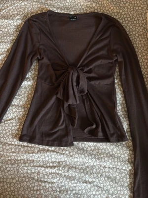 Brun omlottopp från Gina Tricot - Säljer en brun omlottopp från Gina Tricot i mjukt material. Toppen har långa ärmar, v-ringning och knyt framtill som ger en snygg och trendig look. Perfekt att styla med jeans eller kjol för en cool vibe. 💘