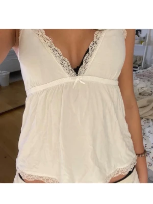 Vit spetskantad nattlinne - Säljer ett vitt nattlinne med tunna axelband och v-ringning. Nattlinnet har dekorativa spetsdetaljer längs halsringningen och nederkanten samt en liten rosett framtill. Materialet är mjukt och lätt, perfekt för att chilla hemma.