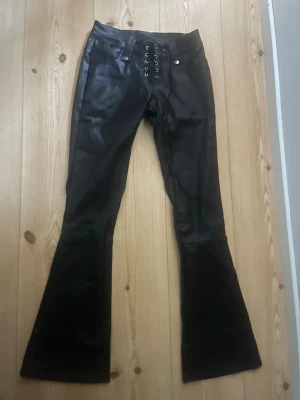 Svarta bootcut skinnbyxor med snörning - Snygga svarta bootcut byxor i syntetmaterial med cool snörning framtill i midjan. Byxorna har fickor både fram och bak samt vida ben som ger en retrovibe. Perfekta för dig som vill sticka ut med en edgy look.