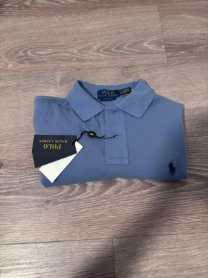 Blå pikétröja från Polo Ralph Lauren - Snygg blå pikétröja från Polo Ralph Lauren med klassisk passform och ikonisk logga broderad på bröstet. Tröjan har krage och knappar framtill, perfekt för en stilren look. Materialet är mjukt och bekvämt bomull.