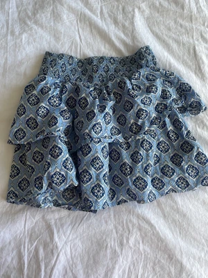 Blå mönstrad volangkjol från Gina Tricot - Säljer en kort blå kjol från Gina Tricot med snyggt mönster och volangdetaljer. Kjolen har smockad midja och är tillverkad i ett lätt bomullstyg som känns skönt mot huden. Perfekt för dig som gillar en trendig och avslappnad stil.