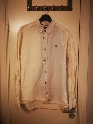 Vit linneskjorta från Henri-Lloyd  - Säljer en stilren vit linneskjorta med klassisk krage och knappar framtill. Skjortan har ett diskret Henri-Lloyd-logo broderat på bröstet och är långärmad. Perfekt för dig som gillar enkel och clean stil.