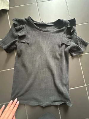 Svart ribbad topp med volangdetaljer - En svart ribbad topp med korta ärmar och volangdetaljer på axlarna. Rund halsringning och stretchigt material som ger en bekväm passform. Perfekt för dig som gillar en enkel men ändå trendig look.