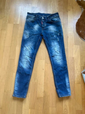 Blå slitna jeans DSQUARED2 (ej äkta) med slim/straight fit - Säljer ett par blå jeans med slim/straight fit och slitningar samt blekta detaljer framtill. Jeansen har klassisk femficksdesign och patch bak på linningen. 