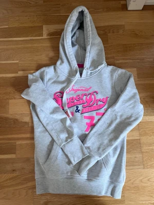 Grå hoodie från Superdry - Grå hoodie från Superdry med rosa tryck och siffran 77 framtill. Klassisk känguruficka och dragsko i huvan. Mjuk bomullsblandning och ribbade muddar vid ärmslut och nederkant. Perfekt för en avslappnad stil.