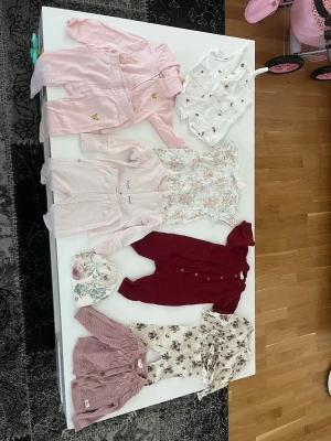 Sött babyklädpaket i pastell och blommigt - Ett paket med babykläder i mjuka pastellfärger och blommiga mönster. Innehåller bodys, sparkdräkter, kofta och mössa i rosa, vinrött, vitt och beige. Plaggen har fina detaljer som volanger, knappar och tryck. Perfekt för dig som gillar gulliga och stilrena babykläder.  Om ni vill endast ha ett plagg, skriv isåfall.