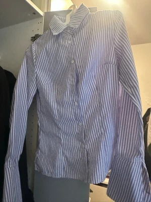 Blårandig skjorta med knappar - Säljer en klassisk blårandig skjorta med vit bas och långa ärmar. Skjortan har knappar framtill och en krage som ger ett stilrent intryck. Perfekt för dig som gillar en tidlös look och vill ha något som funkar till många outfits.