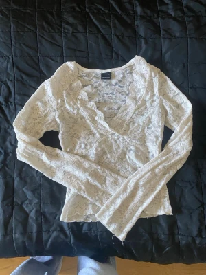 Vit omlottopp i spets från Gina Tricot - Säljer en vit långärmad omlottopp från Gina Tricot i spetsmaterial. Toppen har ett vackert blommigt mönster och en feminin v-ringning. Perfekt för att skapa en romantisk look.