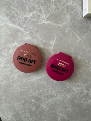 Max Factor Pop Art cheek paint  - Två Max Factor Pop Art cheek paint i rund plastförpackning. Ena är brunrosa och den andra är mörkrosa, båda med svart text på framsidan. Perfekt för att ge kinderna färg och glow. Smidig att ta med i väskan och enkel att applicera.  Obs! Kolla sista bilden, ej använt utan blivit när man öppnat/stängt 