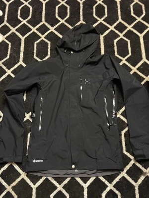 Svart Haglöfs Gore-Tex regnjacka - Svart regnjacka från Haglöfs med Gore-Tex-material, justerbar huva och två stora dragkedjeförsedda fickor på framsidan. Jackan har diskret logotyp på bröstet och är perfekt för att hålla dig torr under regniga dagar.