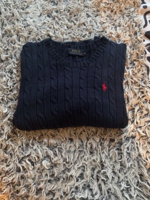 Mörkblå kabelstickad tröja Polo Ralph Lauren - Säljer en mörkblå kabelstickad tröja från Polo Ralph Lauren med klassisk rund halsringning och röd broderad logotyp på bröstet. Den är i fint skick säljer då den inte kommer till användning längre 🤗