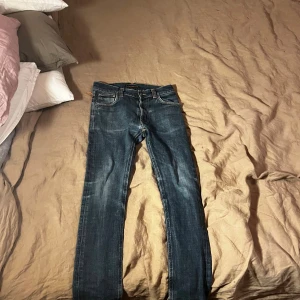 Mörkblå Nudie Jeans - Säljer ett par mörkblå jeans från Nudie Jeans med klassisk femficksmodell och kontrastsömmar. Jeansen är slim och är tillverkade i denim med snygg slitning framtill. Perfekta för dig som gillar en tidlös och avslappnad stil.