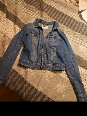 Blå jeansjacka från Gina Tricot - Snygg blå jeansjacka från Gina Tricot med klassisk design, knappar framtill och två bröstfickor med lock. Jackan har en rak passform, krage och långa ärmar. Perfekt att slänga över en t-shirt för en cool look. Materialet är denim med tydliga sömmar.
