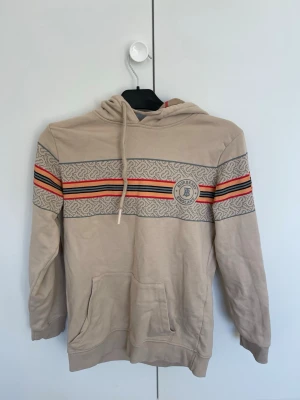 Beige Burberry hoodie med mönster - Snygg beige hoodie från Burberry med klassiskt mönsterband över bröstet och armarna i grått, rött, svart och orange. Stor Burberry-logga på bröstet och typisk rutig insida i huvan. Mjuk bomullskänsla och stor känguruficka framtill.  Den är i M men sitter mer som en S/XS