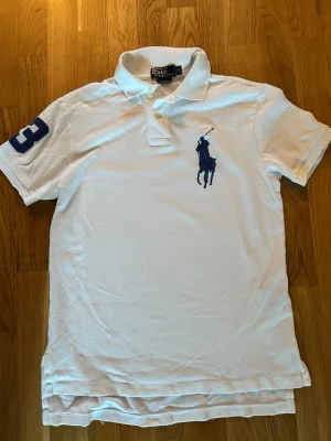 Ralph lauren big pony vintage - Vit Polo Ralph Lauren big pony pikétröja i klassisk och stilren design. Vit färg med mörkblå ikonisk logga på bröstet och en stor “3” på ärmen som ger en sportig look. Perfekt både till vardags och mer uppklädda tillfällen. Tillverkad i klassisk bomullspiké som är skön att bära. Tröjan är i bra använt skick – något skrynklig på bilderna men hel och fin. Den har även några små upprivningar i armar och nacke se på bilder. Passar bra till jeans, chinos eller shorts.