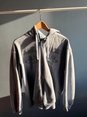 Grå zip hoodie med tryck - Grå hoodie med dragkedja från Bling Lucid. På bröstet finns ett tryck med två änglar och texten 'BLING LUCID'. Hoodien har huva med dragsko och ribbade muddar vid ärmar och nederkant. Perfekt för en chill och avslappnad stil.