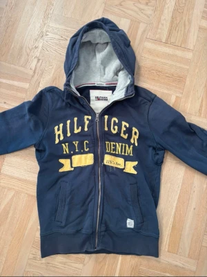 Marinblå Hilfiger Denim hoodie - Snygg marinblå vintage hoodie från Hilfiger Denim med dragkedja och huva. Stora gula bokstäver och detaljer framtill med texten 'HILFIGER N.Y.C DENIM 115.7th Ave.'. Mjuk insida och två fickor fram. Perfekt för en avslappnad streetwear-look. (Unisex)