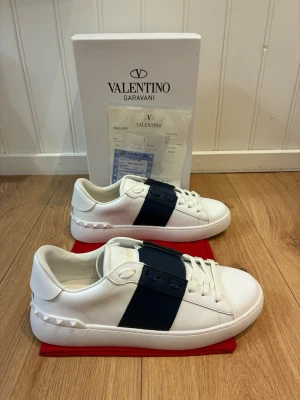 Valentino Garavani open sneakers  - Snygga vita sneakers från Valentino Garavani med bred mörkblå rem över vristen och ikoniska nitar på hälen. Skorna är i läder och har klassisk låg profil med vita snören och diskret guldtext på plösen. Perfekta för dig som gillar stilrena och exklusiva detaljer.