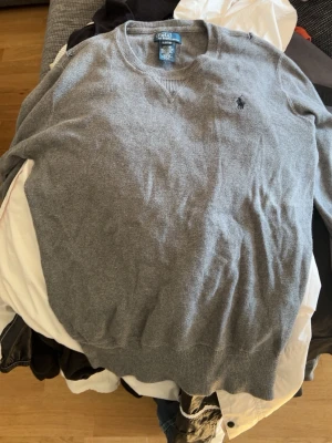 Grå tröja från Polo Ralph Lauren - Klassisk grå tröja från Polo Ralph Lauren i 100% bomull. Tröjan har rund halsringning, ribbade muddar och den ikoniska broderade loggan på bröstet. Perfekt för en stilren och avslappnad look.