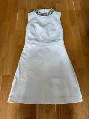 Vit klänning med pärlkrage - Snygg vit klänning med ärmlös design och en dekorerad pärlkrage i silver. Klänningen har en lätt utställd kjol och dragkedja i ryggen. Perfekt för dig som vill sticka ut med eleganta detaljer och stilren look.
