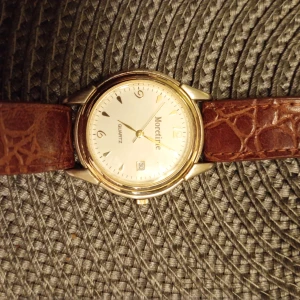 Moretime klocka med brun läderrem - Stilren klocka från Moretime med rund, guldig boett och vit urtavla. Brunt läderarmband med krokopräglat mönster. Urtavlan har datumvisning och tydliga siffror. Quartz-urverk och baksida i rostfritt stål. Nytt batteri behövs.