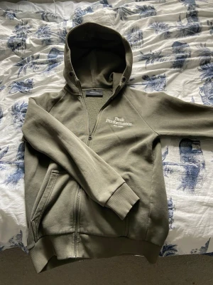Olivgrön hoodie från Peak Performance - Olivgrön hoodie från Peak Performance med dragkedja och broderad logga på bröstet. Mjuk fleece på insidan, ribbade muddar och två sidofickor. Riktigt bra skick och inga skador. 