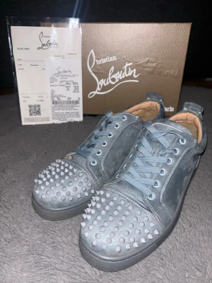 Gråa Christian Louboutin sneakers - Snygga gråa sneakers från Christian Louboutin i mocka med ikoniska nitar på tån och klassisk röd sula. Skorna har blågrå skosnören och en låg silhuett som ger en cool och edgy vibe. Perfekta för dig som vill sticka ut med exklusiva detaljer. OBS!!! en spik saknas. Pris kan diskuteras vid snabb affär!!!
