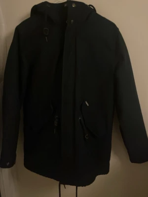 Mörkblå parkajacka från Carhartt M - Säljer en svart parkajacka från Carhartt i storlek M. Jackan har huva, snörning i midjan och nederkant, stora fickor framtill och dragkedja med tryckknappar. 