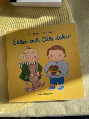 Ellen och Olle leker av Kruusval - Charmig barnbok med hårda sidor, illustrerad av Catarina Kruusval. Boken har ett gult omslag med tecknade figurer och innehåller färgglada bilder på saker som ballong och skor. Perfekt för små barn som vill lära sig ord och upptäcka världen.