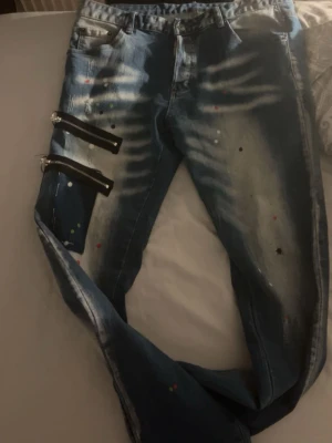 Blå jeans med dragkedjor och färgstänk - Säljer ett par blå jeans med coola dragkedjor på ena benet och unika färgstänk över hela byxan. Jeansen har en tvättad look med ljusa partier och två bakfickor. Perfekt för dig som vill sticka ut med din stil.