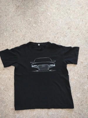Svart t-shirt med Audi-motiv - Cool svart t-shirt med ett vitt tryck av en Audi-bil på bröstet. Klassisk rund hals och korta ärmar, perfekt för dig som gillar bilar och vill ha en enkel men snygg stil. Materialet känns mjukt och bekvämt, troligen bomull.