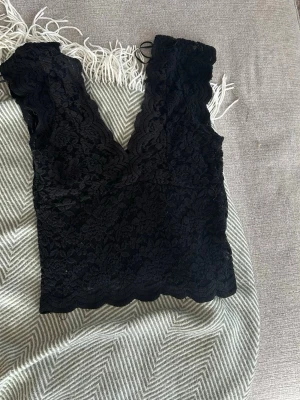 Svart spetsig croppad topp - Säljer en svart croppad topp med v-ringning och breda axelband. Toppen är helt täckt i vacker spets med scallop-kant och har en feminin och elegant look. Perfekt för att styla med jeans eller kjol.