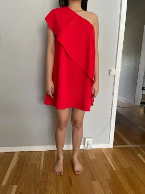 Röd oneshoulder kortklänning - En snygg röd kortklänning med oneshoulder-design och volangdetalj som ger ett coolt och trendigt uttryck. Klänningen har en lös passform. Det står L på etiketten men den passar snarare till M. Hon som är på bild är 174/65.